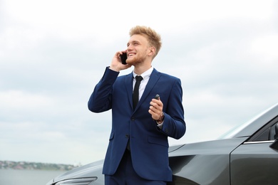 Photo of Young businessman with key talking on phone near car outdoors