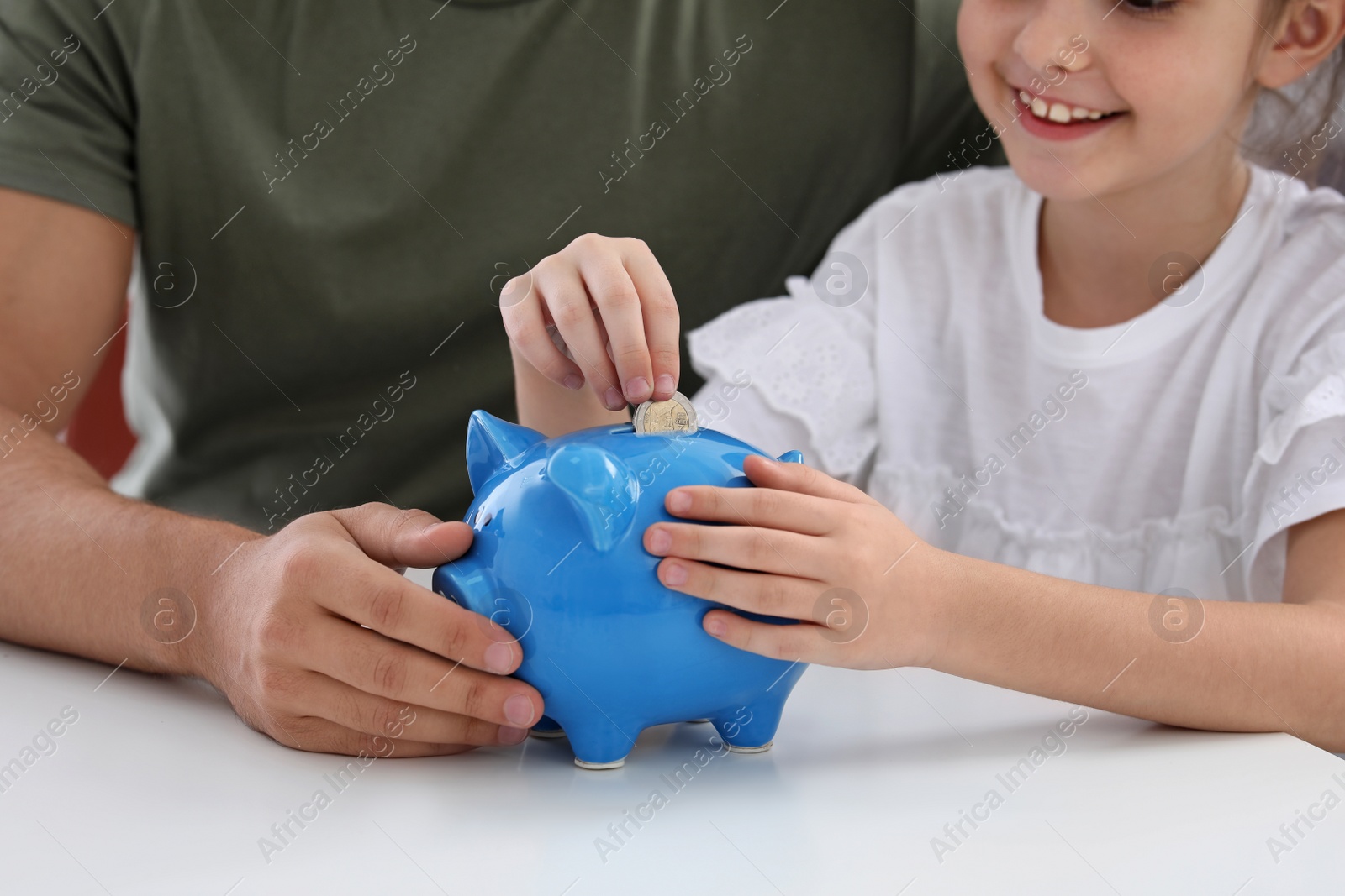 Photo of Little girl with her father putting coin into piggy bank at table, closeup