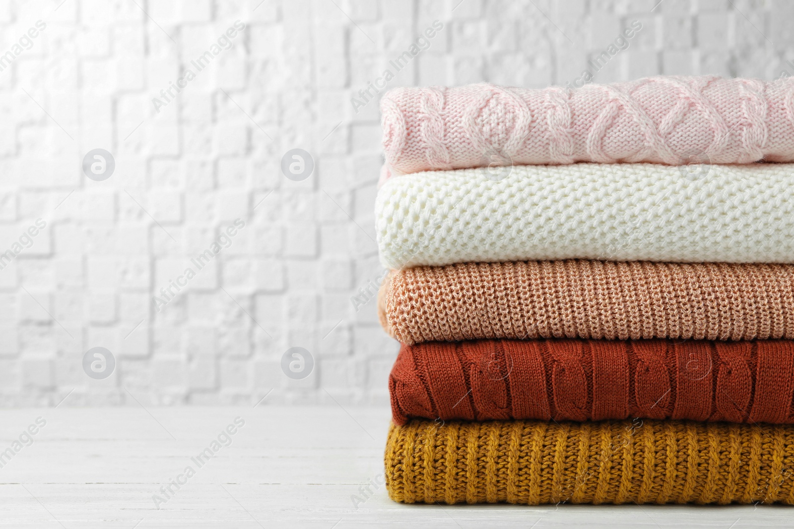 Photo of Stack of warm clothes on white wooden table against textured wall, space for text. Autumn season