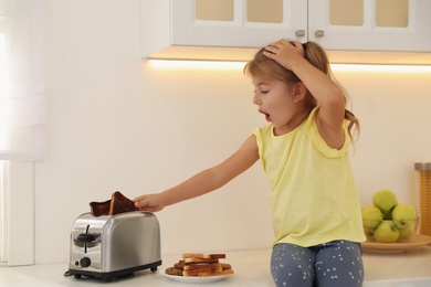 Emotional girl taking slice of burnt bread from toaster in kitchen