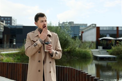 Sleepy man with cup of coffee yawning near river outdoors. Space for text