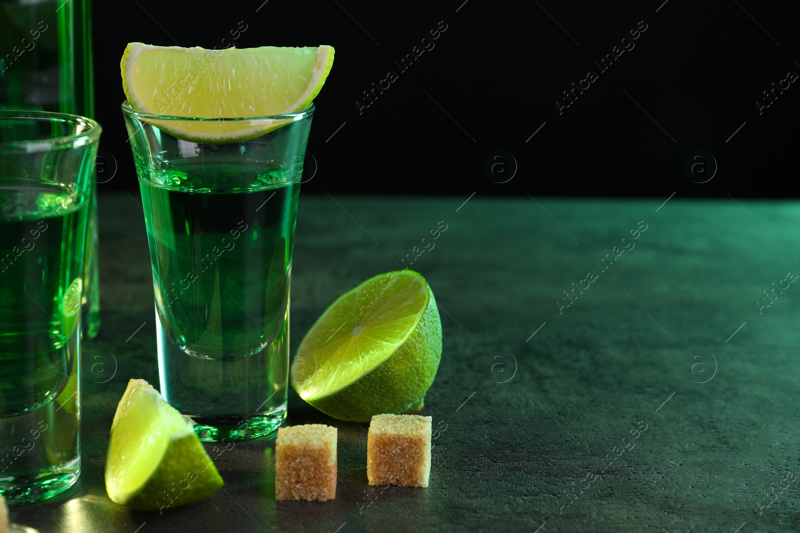 Photo of Absinthe in shot glasses, lime and brown sugar cubes on gray textured table against black background, space for text. Alcoholic drink