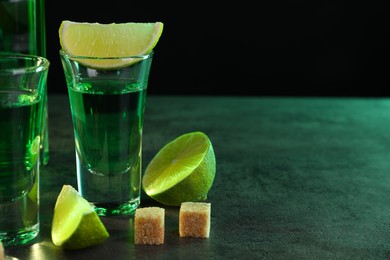 Absinthe in shot glasses, lime and brown sugar cubes on gray textured table against black background, space for text. Alcoholic drink
