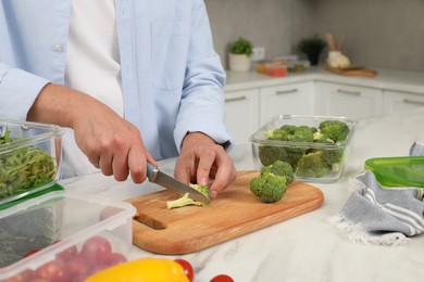 Photo of Man cutting fresh broccoli with knife near containers at white marble table in kitchen, closeup. Food storage