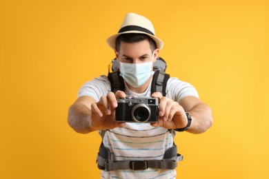 Male tourist in protective mask with travel backpack taking picture against yellow background, focus on camera