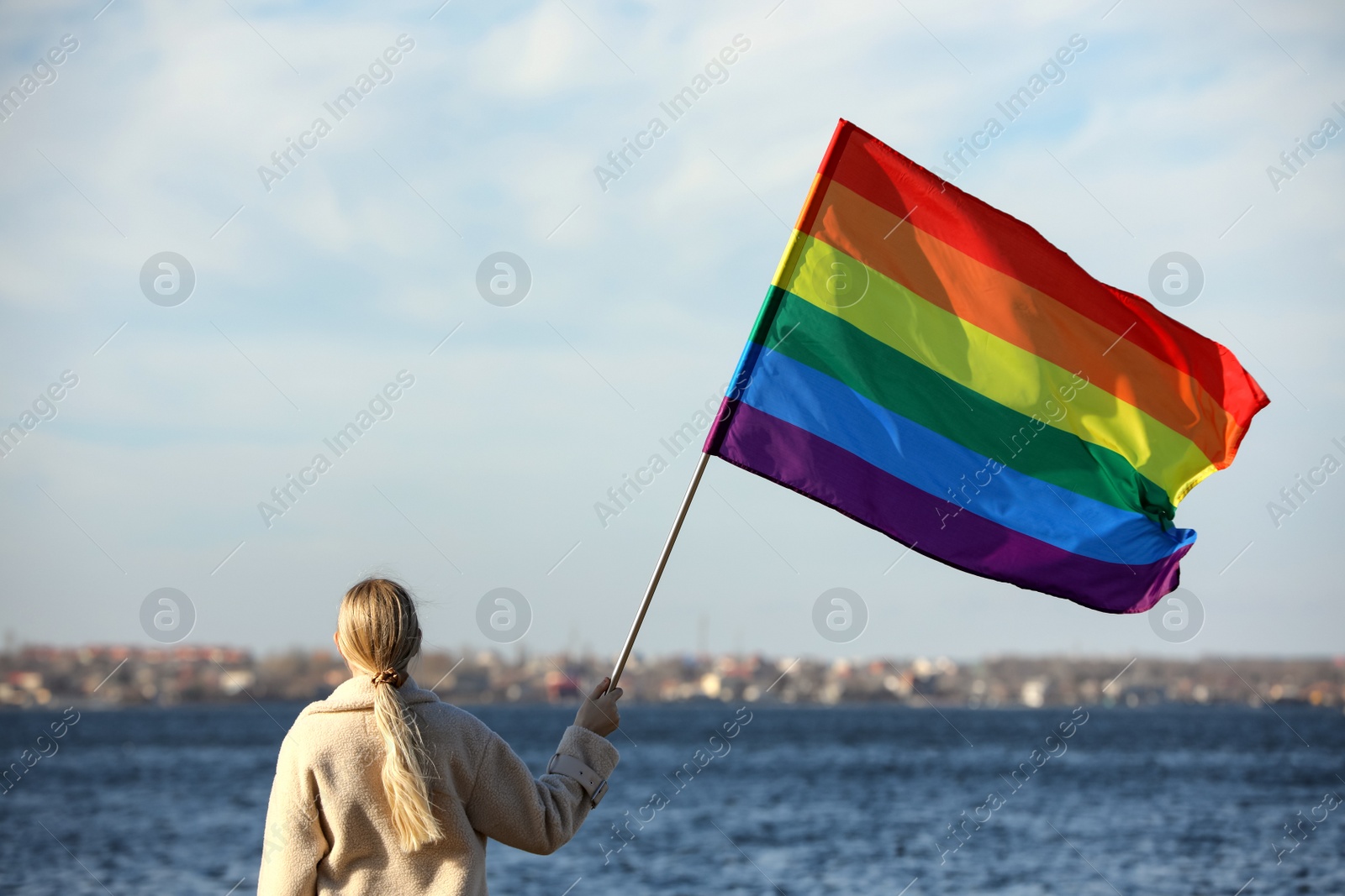 Photo of Woman holding bright LGBT flag near river, back view