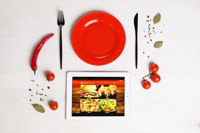 Photo of Modern tablet with open page for online food ordering, scattered vegetables, spices, plate and cutlery on white wooden table, flat lay. Concept of delivery service
