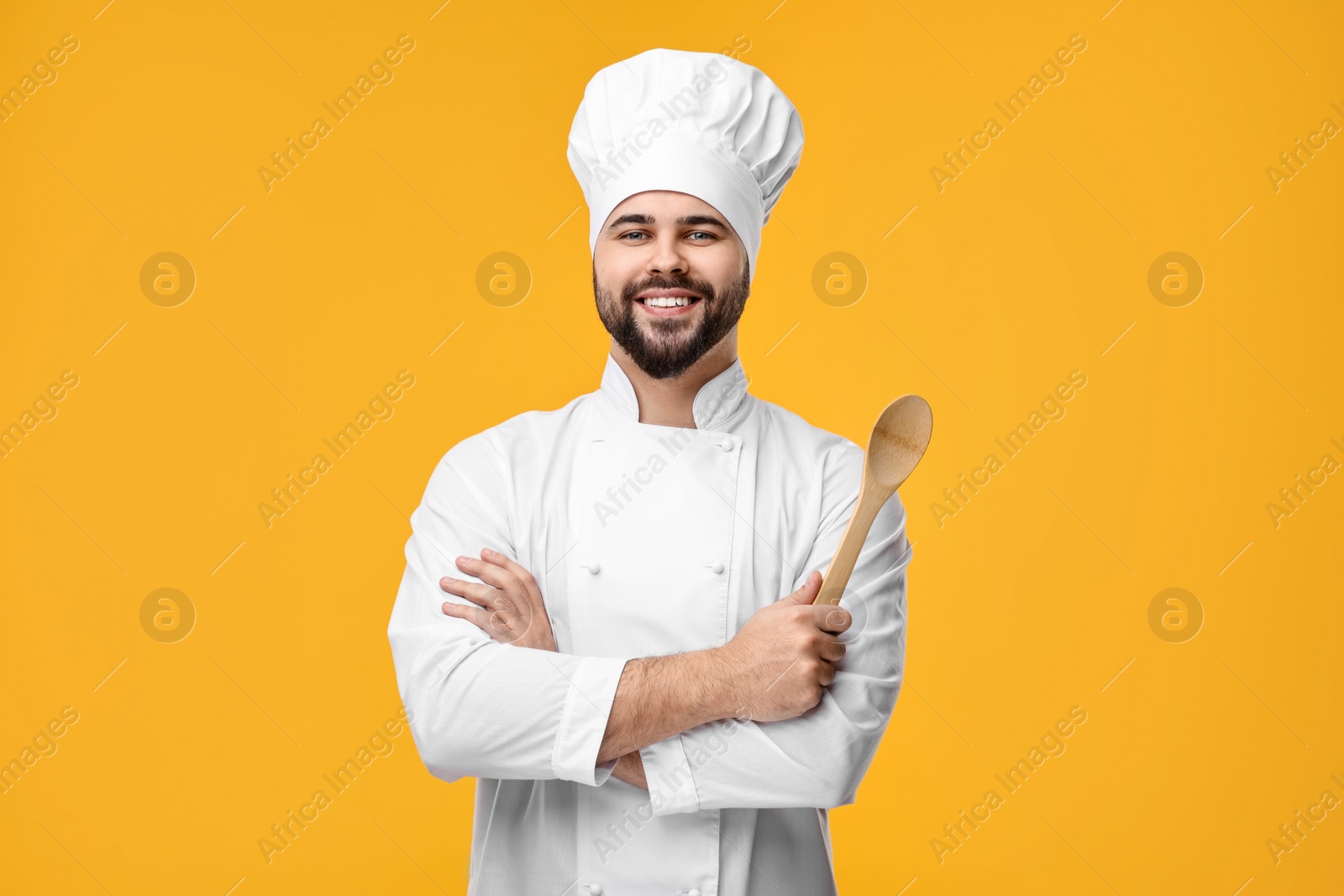 Photo of Happy young chef in uniform holding wooden spoon on orange background