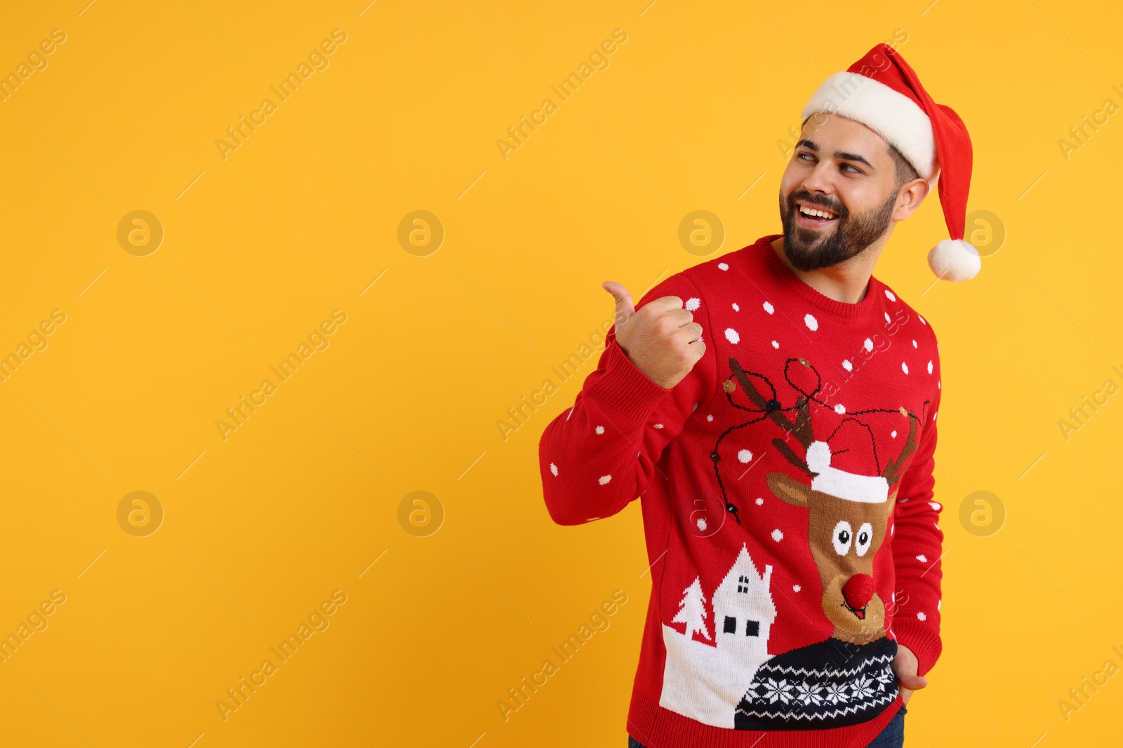 Photo of Happy young man in Christmas sweater and Santa hat pointing at something on orange background. Space for text