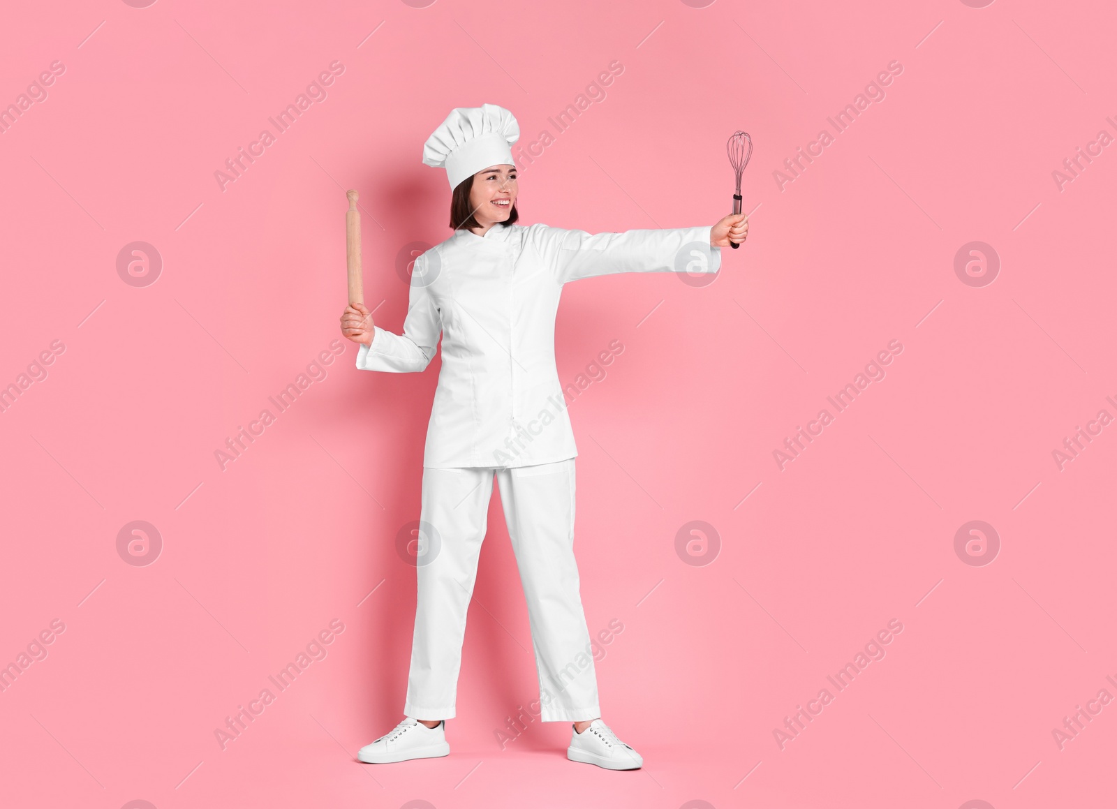 Photo of Happy confectioner with rolling pin and whisk on pink background