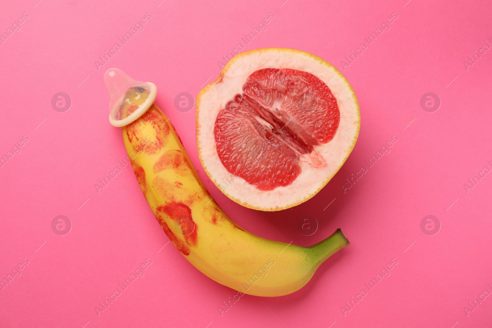 Photo of Half of grapefruit, banana with condom and red lipstick marks on pink background, flat lay. Safe sex concept