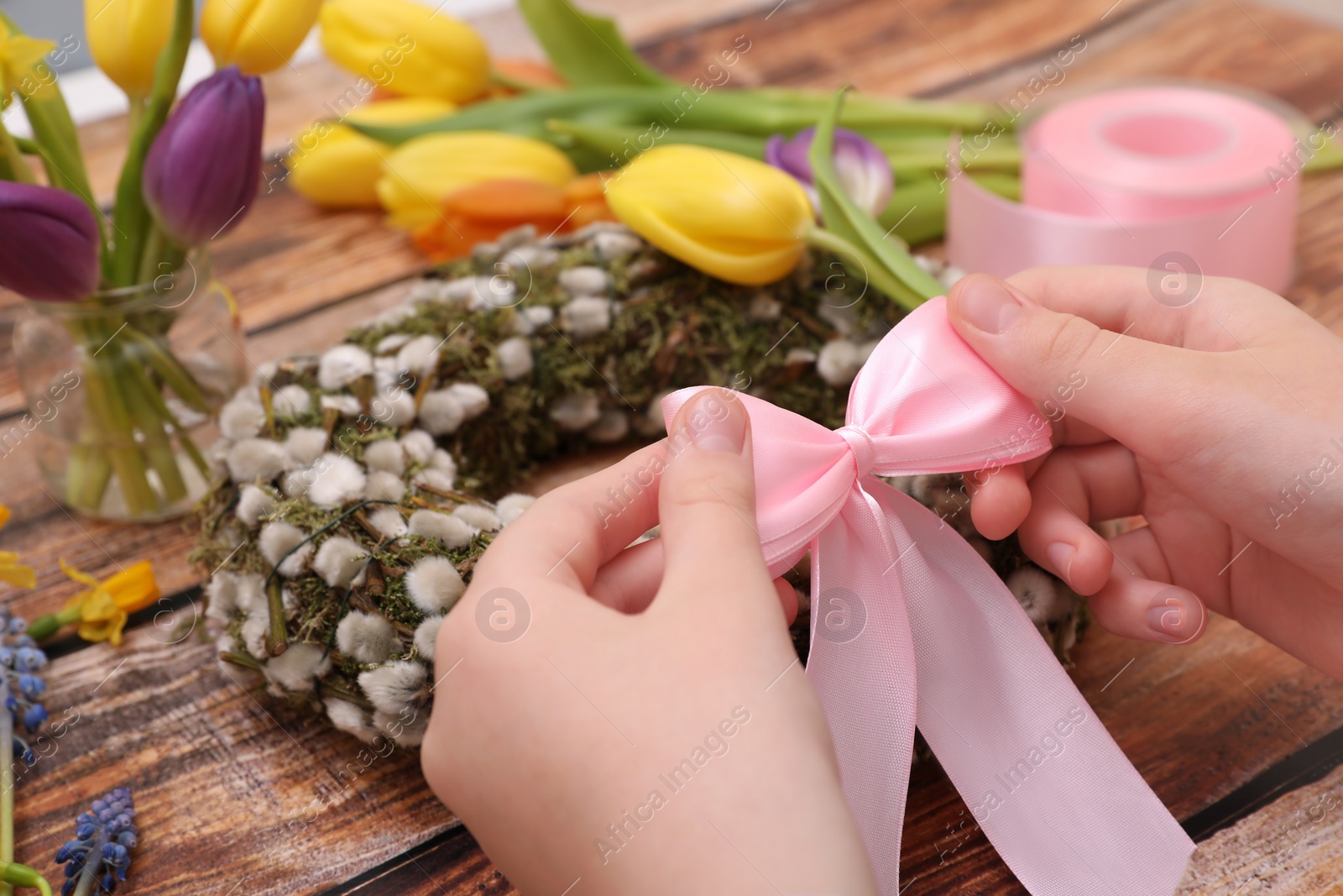 Photo of Woman decorating willow wreath with pink bow at wooden table, closeup