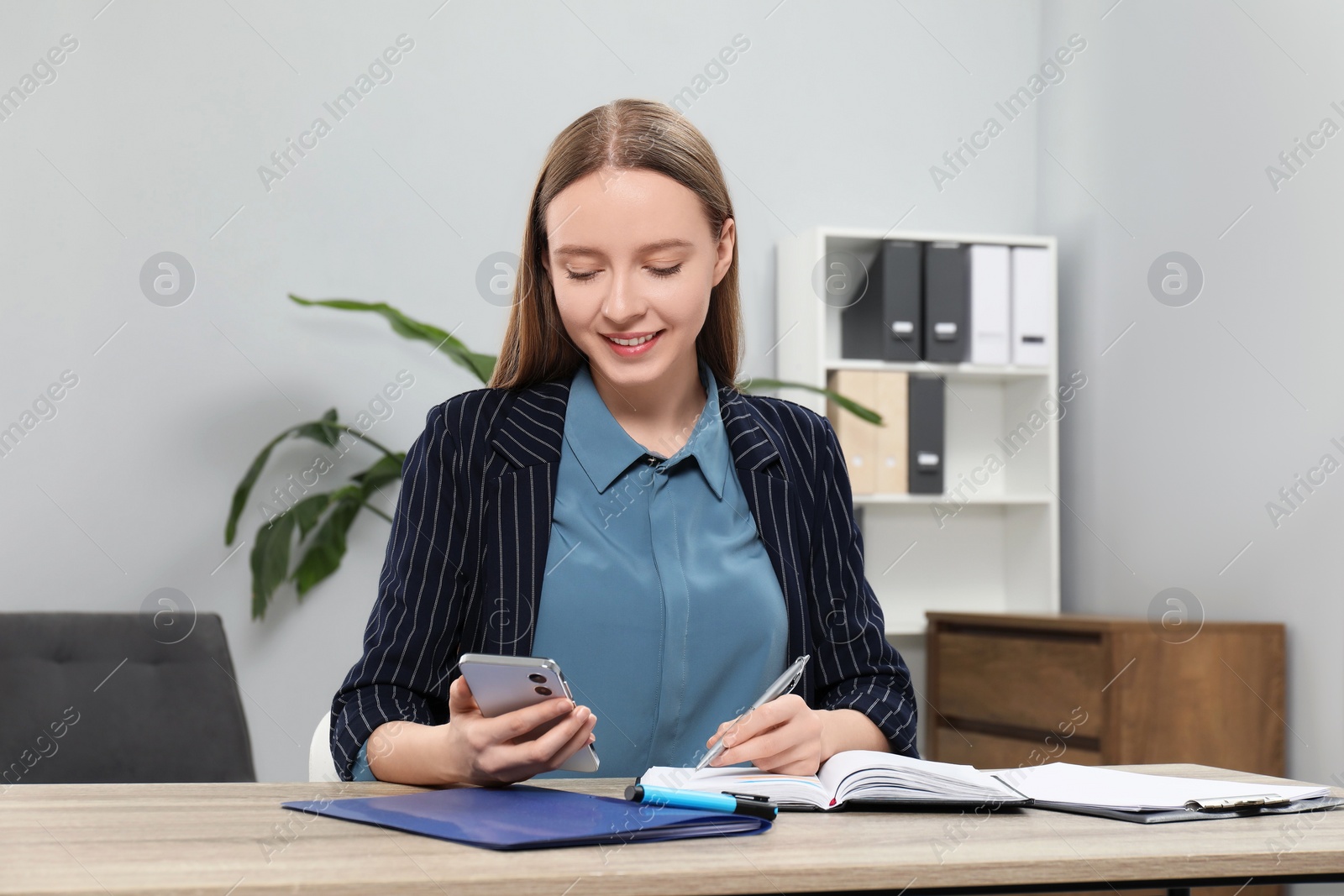 Photo of Woman taking notes while using smartphone at wooden table in office