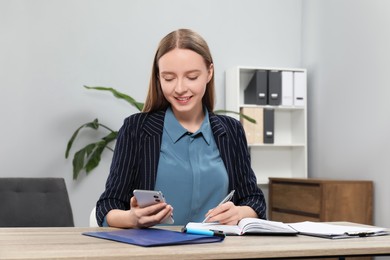 Photo of Woman taking notes while using smartphone at wooden table in office