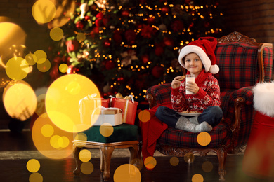 Photo of Little boy in Santa Claus cap with milk and cookies near Christmas gifts at home
