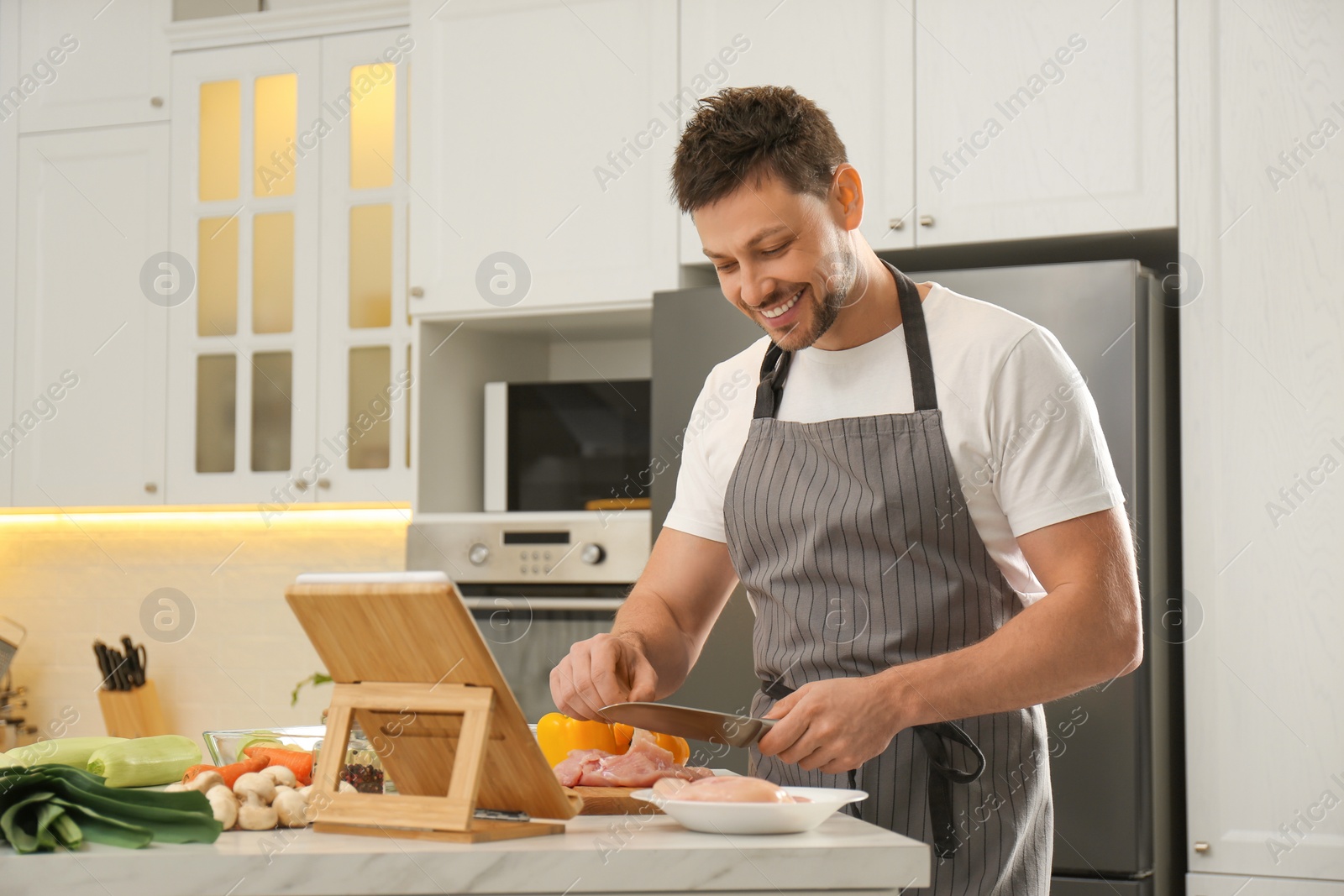 Photo of Man cutting chicken fillet while watching online cooking course via tablet in kitchen