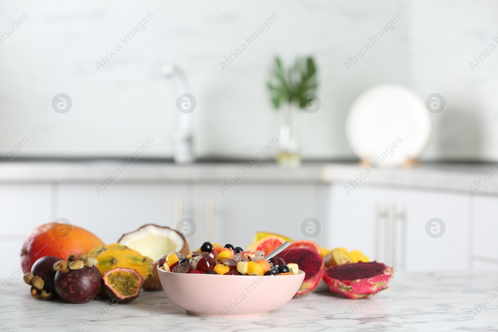 Photo of Delicious exotic fruit salad and ingredients on white marble table in kitchen. Space for text