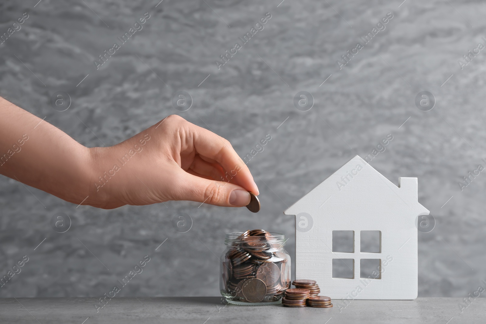 Photo of Woman putting coin into glass jar near house model on table against grey background. Space for text