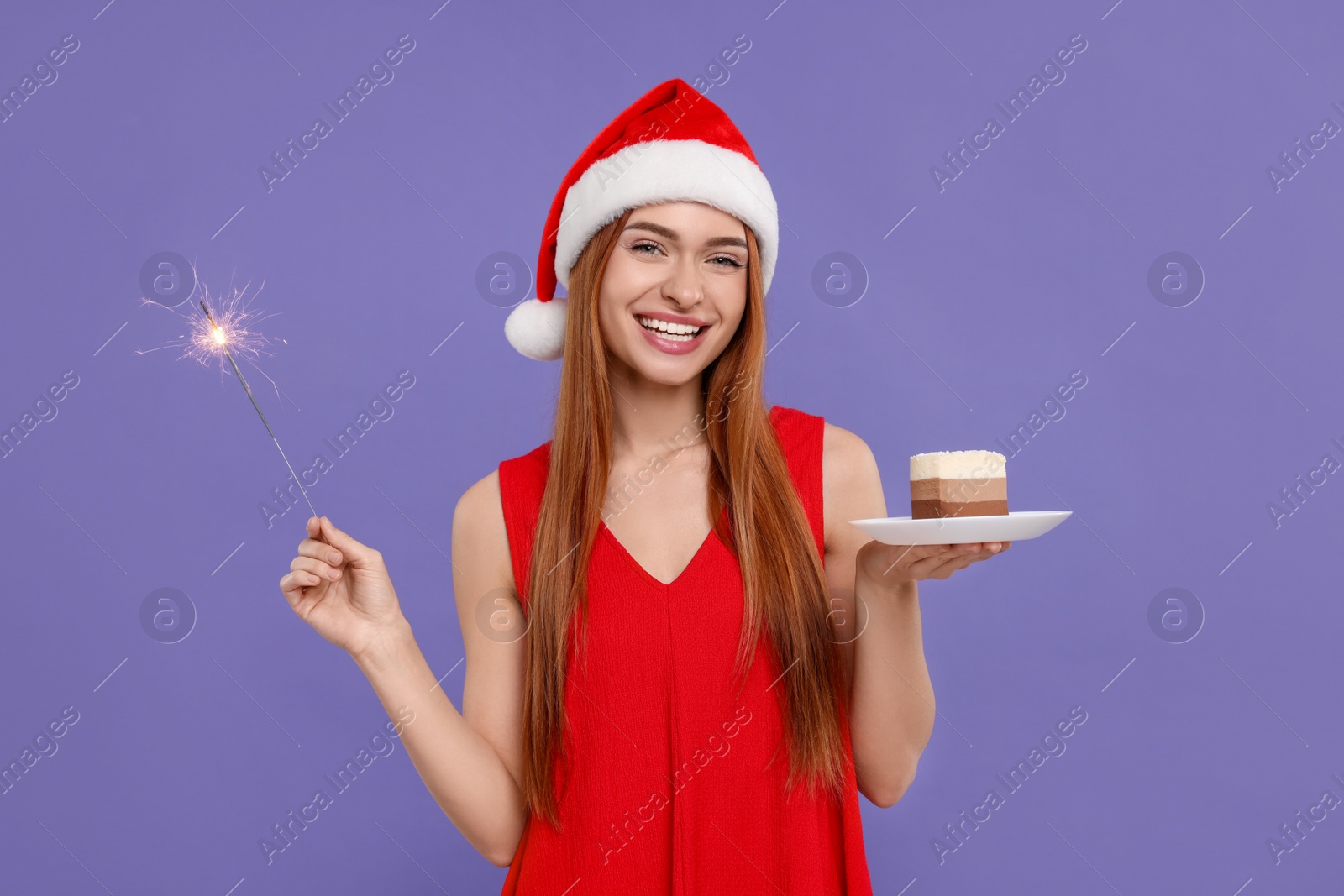 Photo of Young woman in Santa hat with piece of cake and burning sparkler on purple background