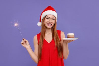Young woman in Santa hat with piece of cake and burning sparkler on purple background