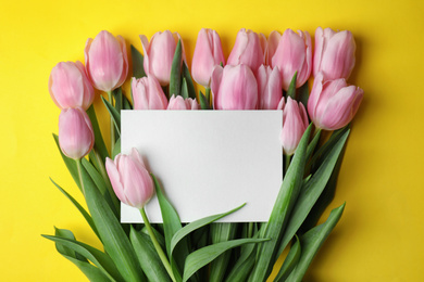 Beautiful pink spring tulips and blank card on yellow background, flat lay. Space for text