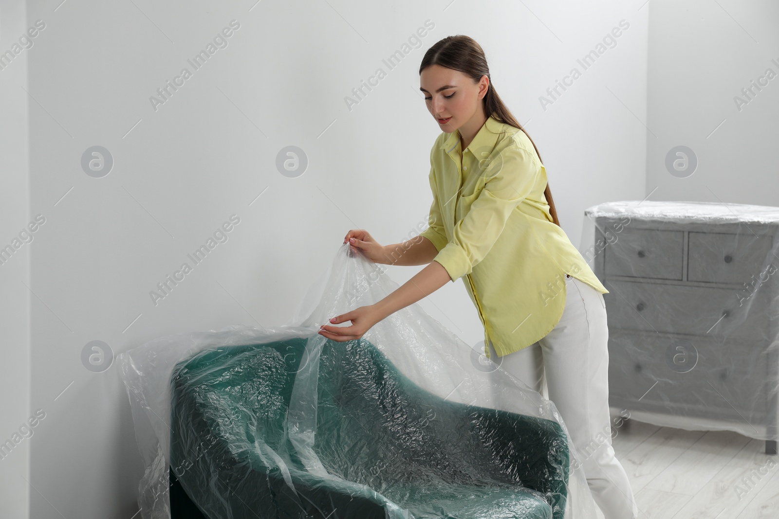 Photo of Young woman putting plastic film away from armchair near light grey wall indoors