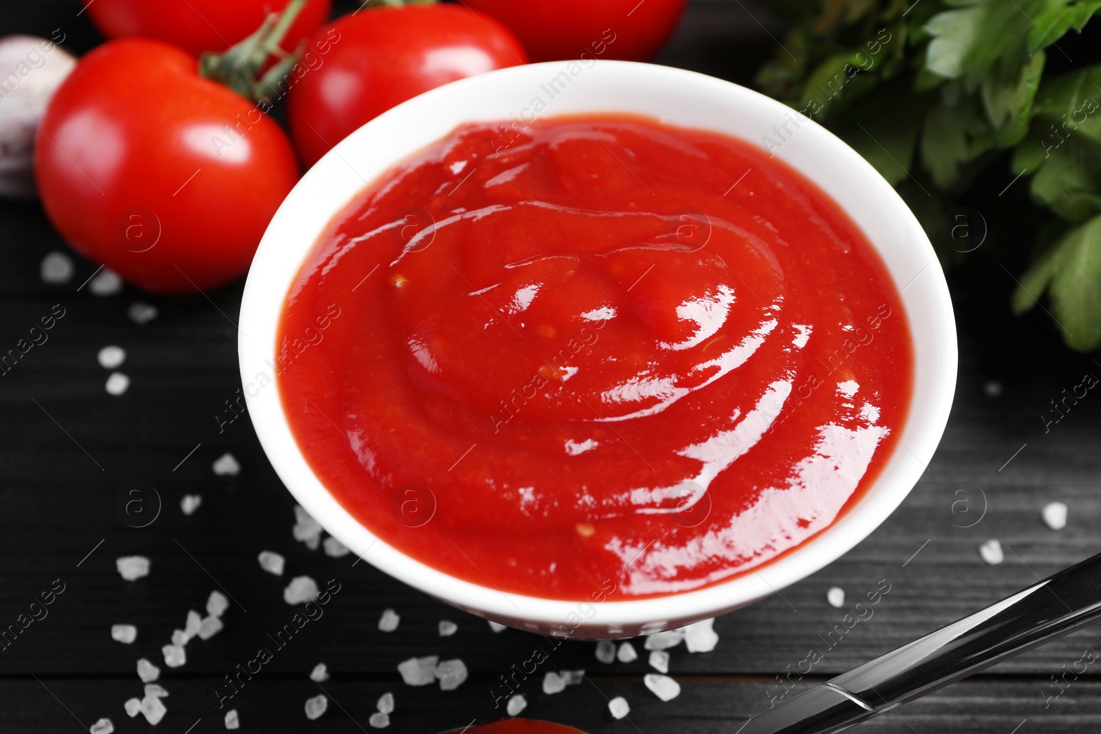 Photo of Delicious ketchup in bowl and salt on black wooden table, closeup. Tomato sauce