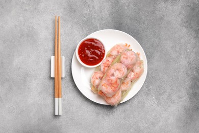 Tasty spring rolls with sauce and chopsticks on grey textured table, flat lay