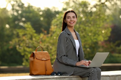Beautiful young woman with stylish backpack and laptop on bench in park