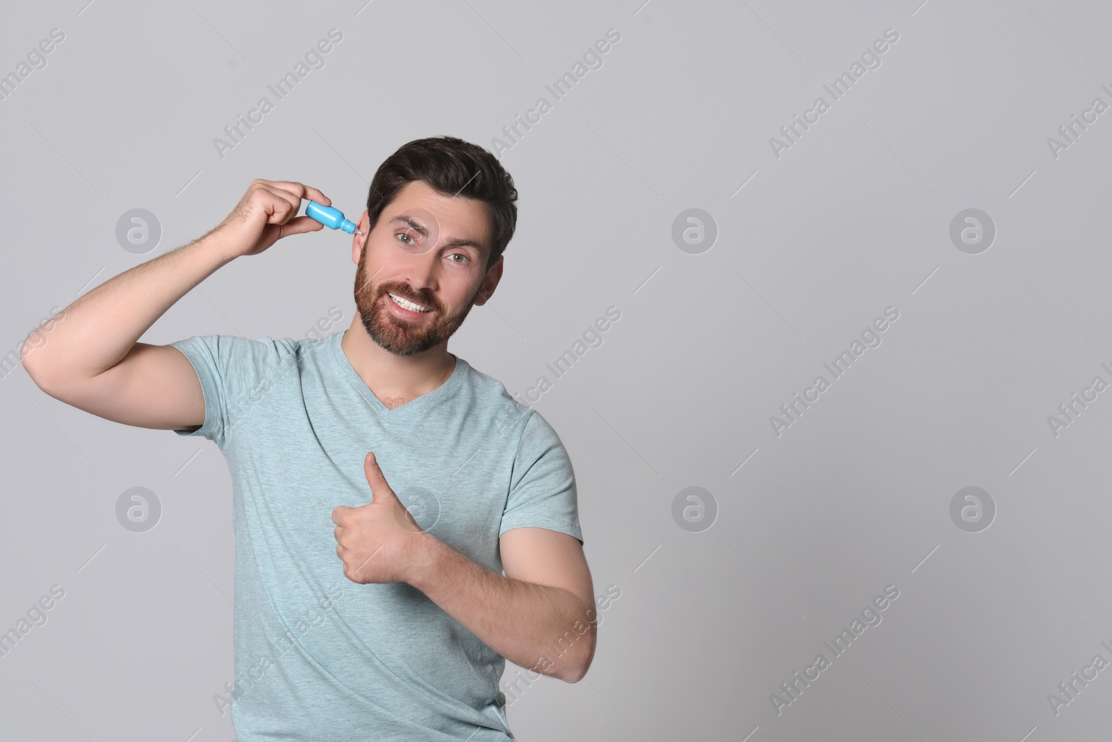 Photo of Man using ear drops and showing thumbs up on grey background, space for text