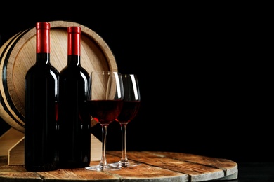 Photo of Glasses with delicious wine, bottles and barrel on table against dark background