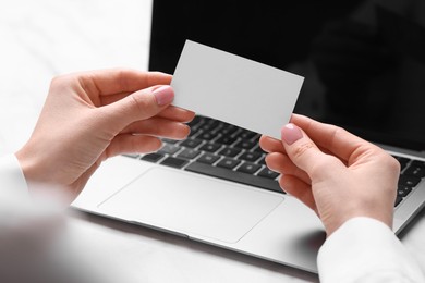 Woman with laptop holding blank business card at white table, closeup. Space for text