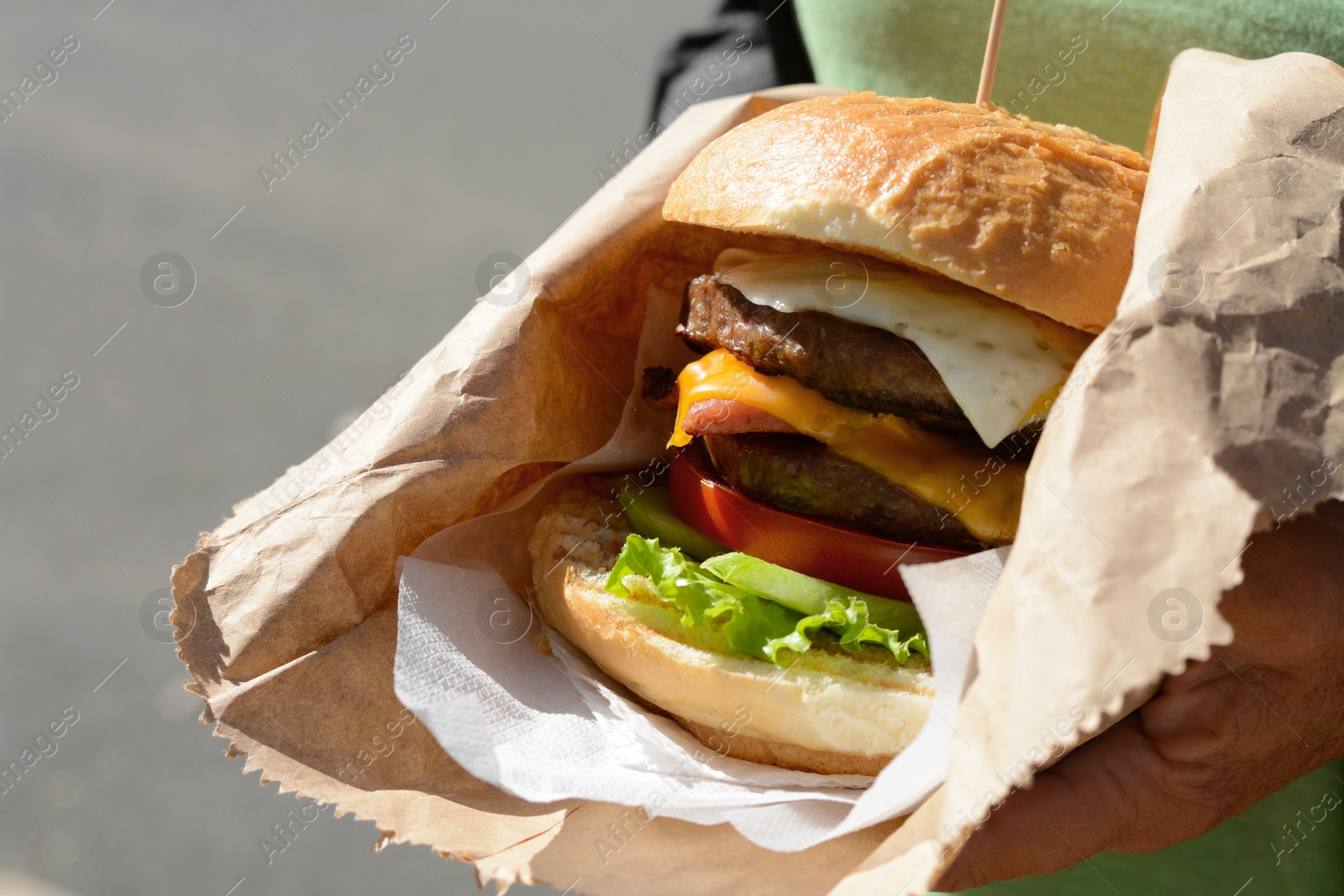 Photo of Woman holding delicious burger in paper wrap on blurred background, closeup