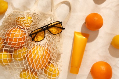 Photo of String bag with sunglasses, fruits and sunscreen on sand, flat lay