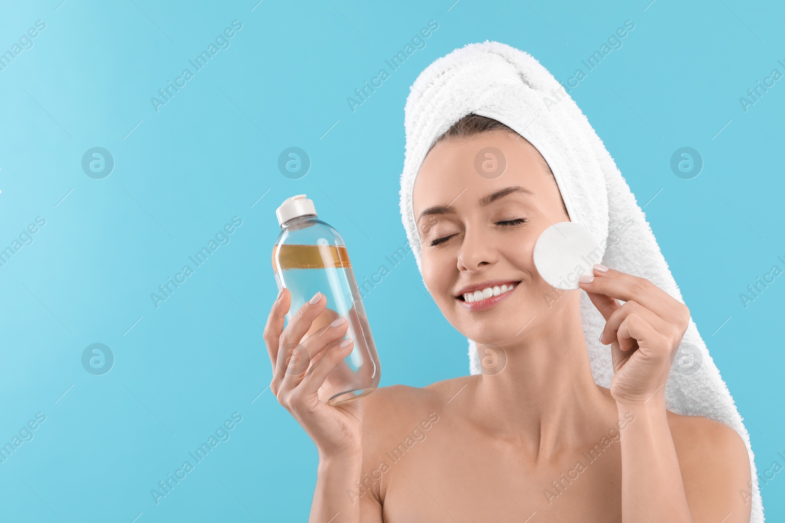 Photo of Smiling woman removing makeup with cotton pad and holding bottle on light blue background. Space for text