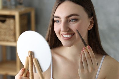 Smiling woman drawing freckles with pen in front of little mirror indoors