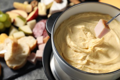 Dipping piece of ham into fondue pot with melted cheese at table with snacks, closeup