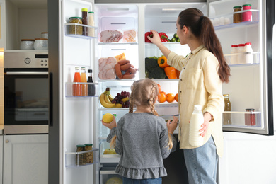 Photo of Young mother and her daughter with products near open refrigerator in kitchen