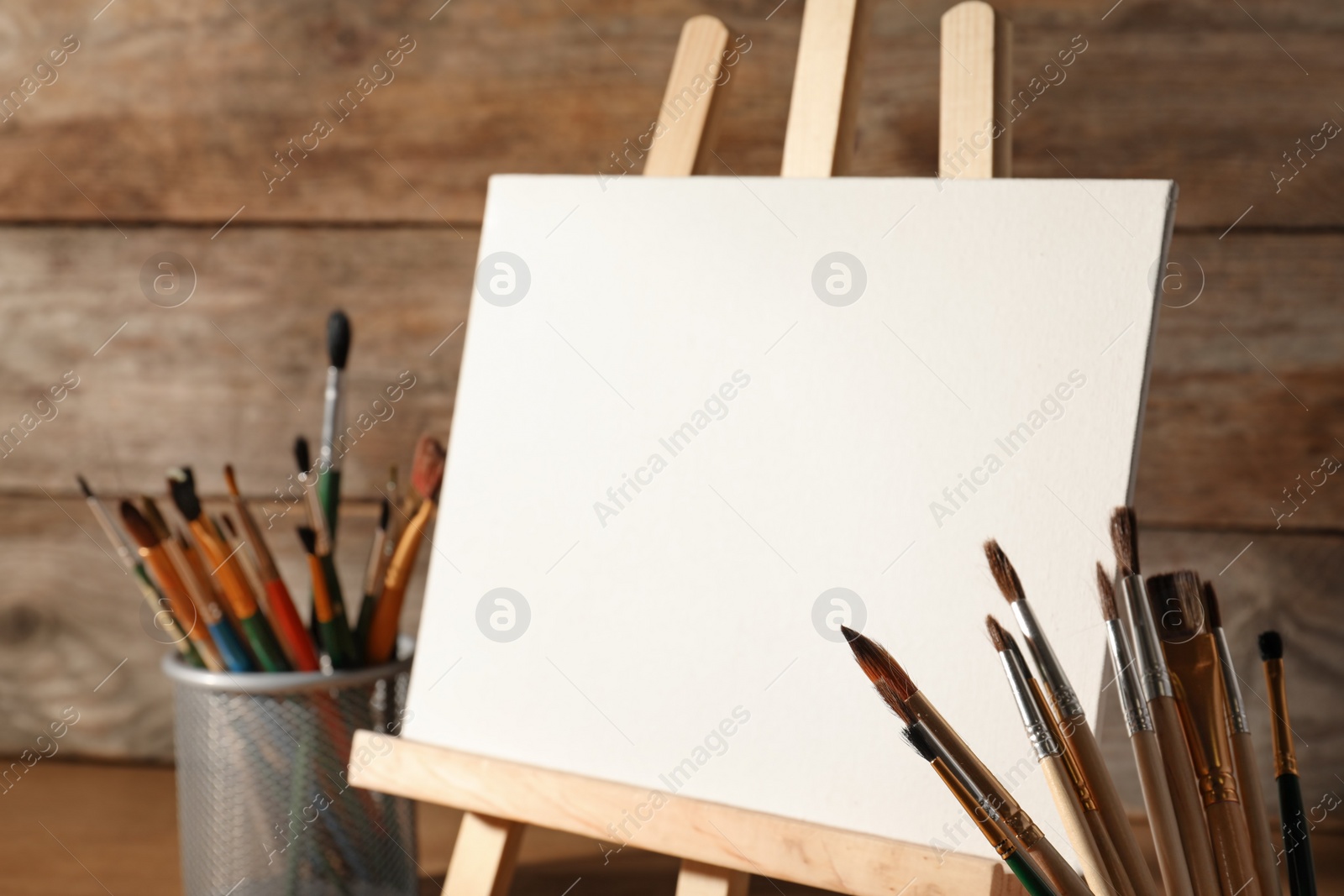 Photo of Easel with blank canvas board and brushes on table near wooden wall. Children's painting