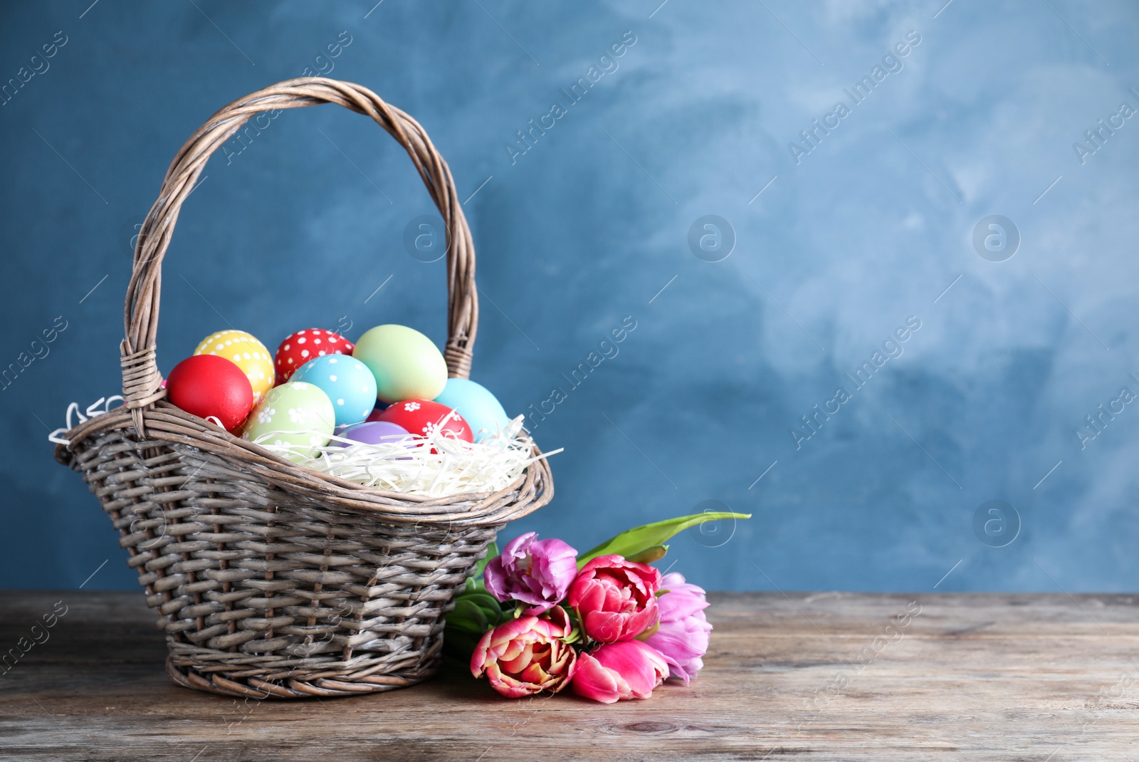 Photo of Wicker basket with bright painted Easter eggs and spring flowers on wooden table against blue background. Space for text