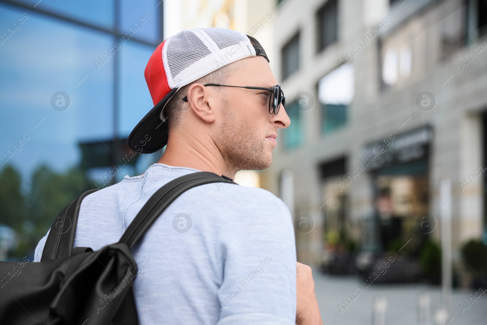 Photo of Handsome young man with stylish sunglasses and backpack near building outdoors, space for text