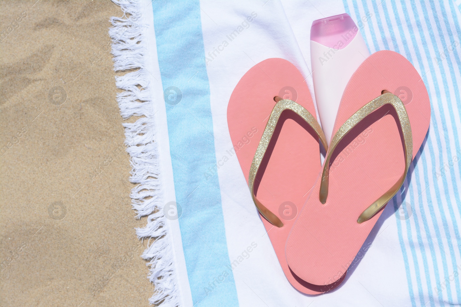 Photo of Striped towel with bottle of sunblock and flip flops on sandy beach, flat lay. Space for text