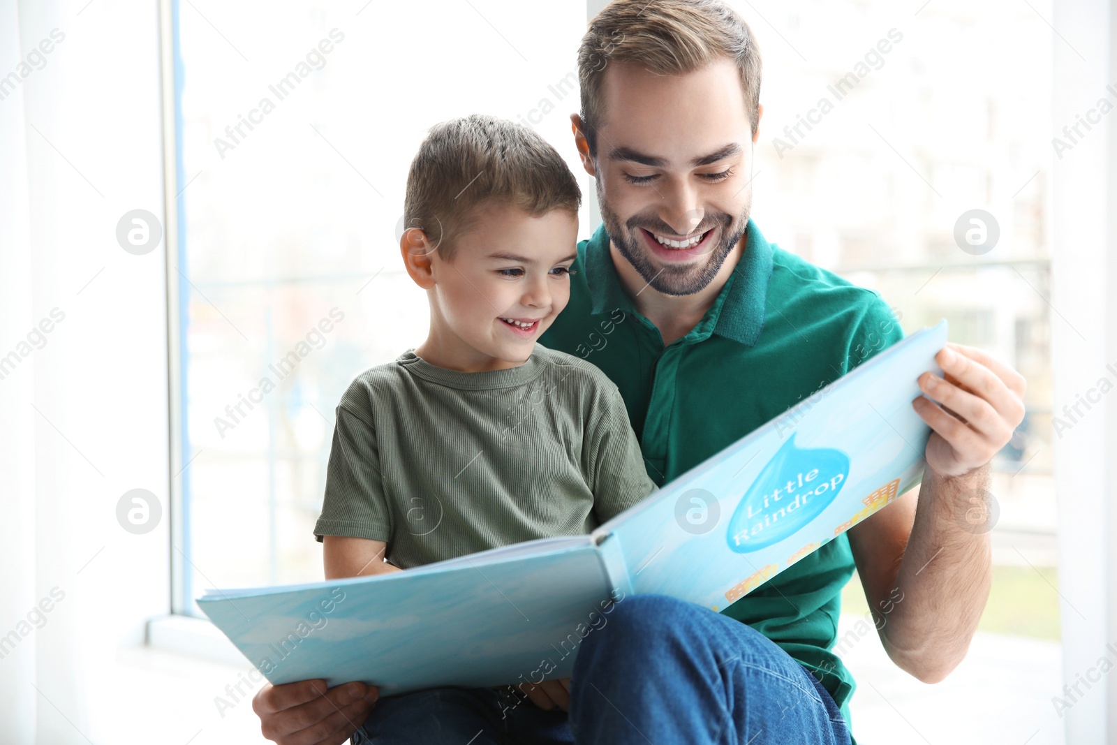 Photo of Young man and his little son reading book near window at home