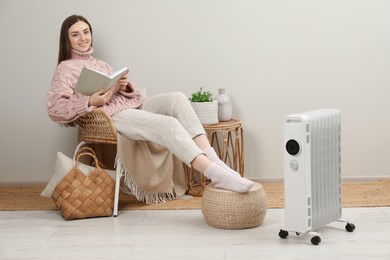 Photo of Woman reading book near modern portable electric heater indoors