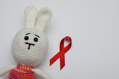 Photo of Cute knitted toy bunny and red ribbon on light grey background, flat lay with space for text. AIDS disease awareness