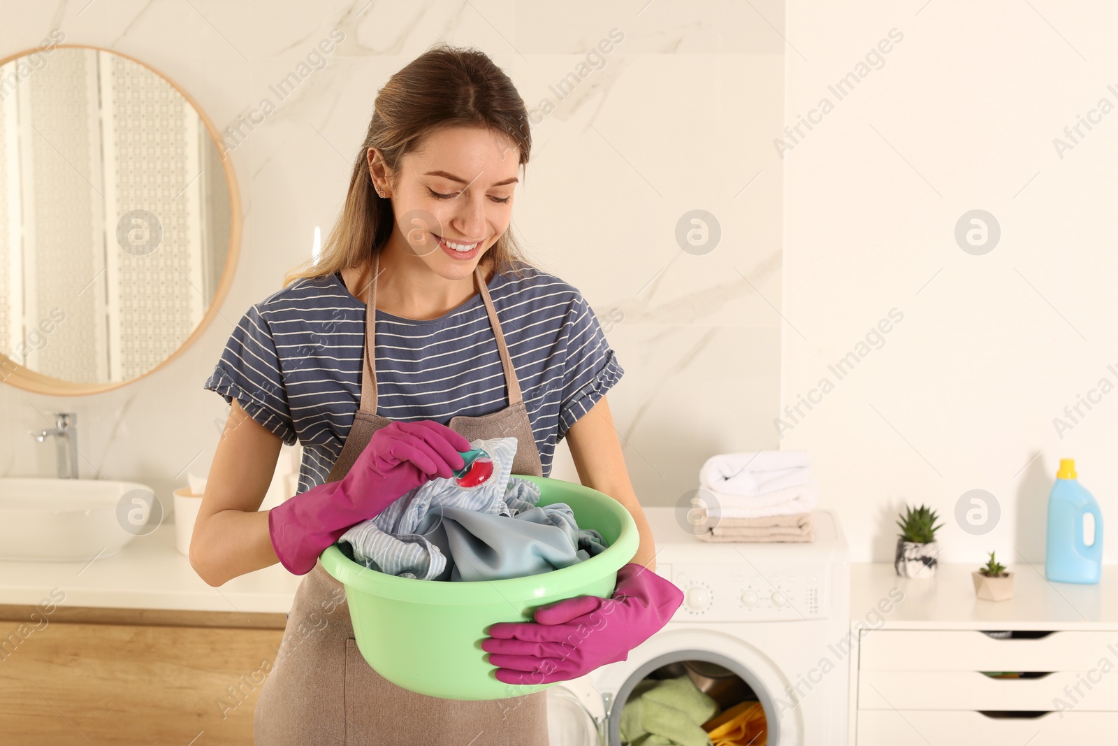 Photo of Woman holding dirty clothes and laundry detergent capsule in bathroom