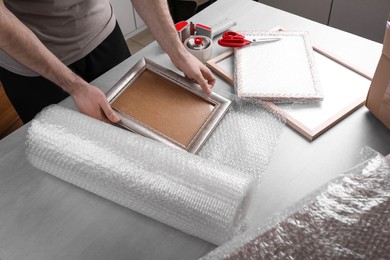 Man covering photo frame with bubble wrap at light grey table, closeup