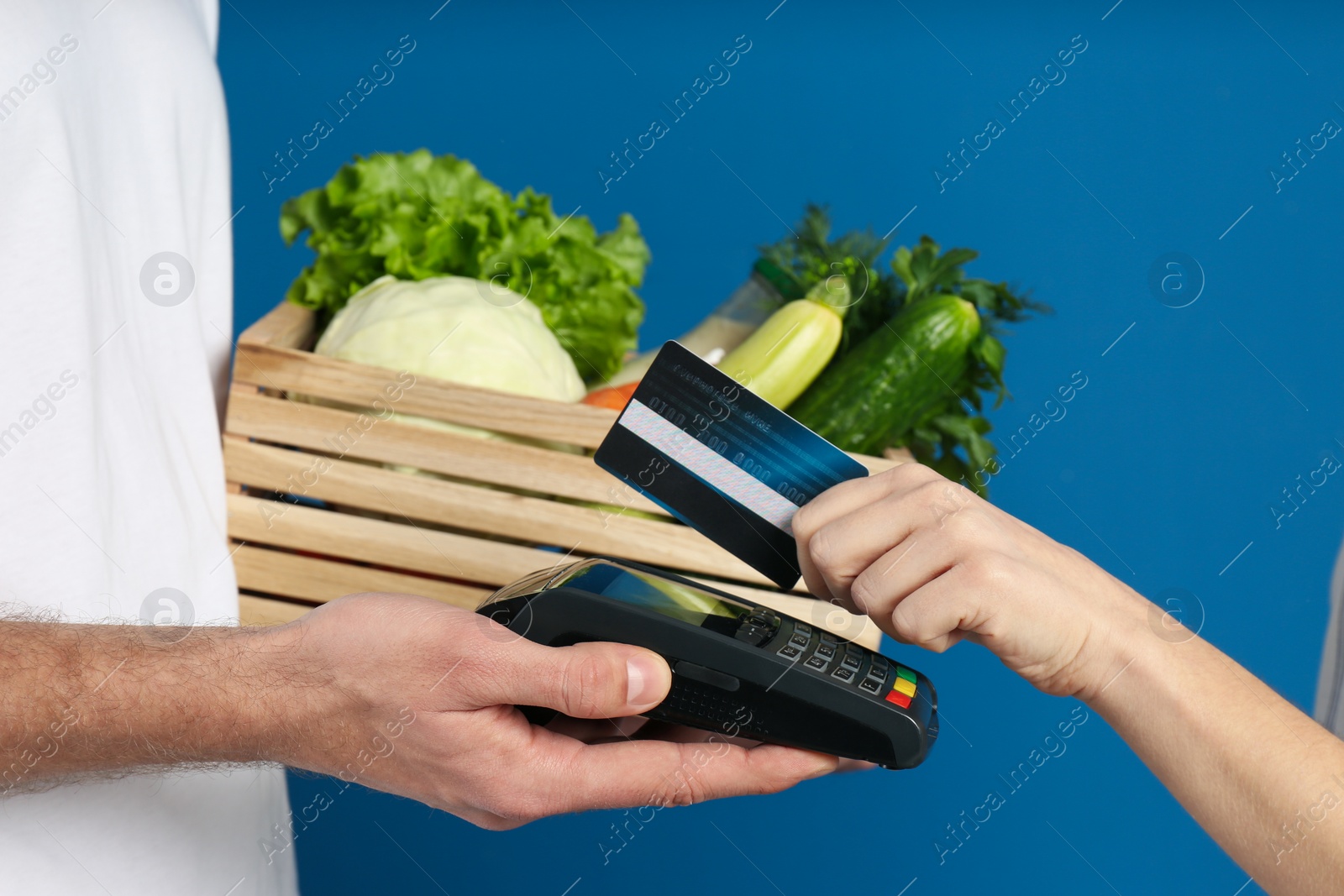Photo of Client paying courier for fresh products with credit card on blue background, closeup. Food delivery service