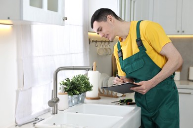 Photo of Smiling plumber with clipboard examining faucet in kitchen