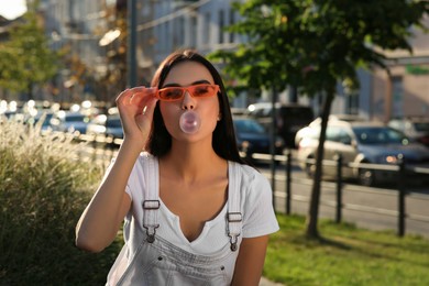 Beautiful woman in sunglasses blowing gum outdoors, space for text